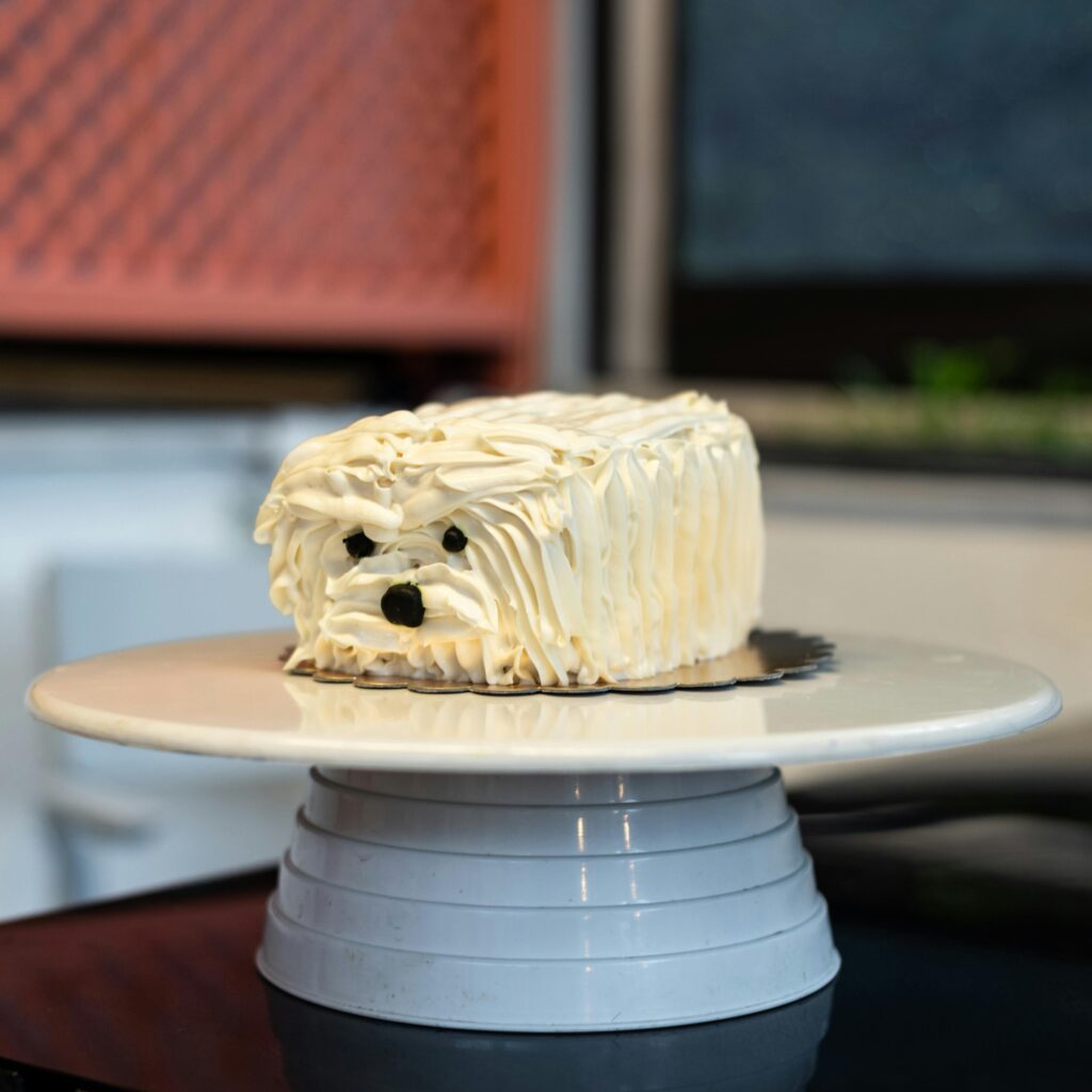 A cute cake sculpted to resemble a fluffy white puppy, displayed indoors on a stand.