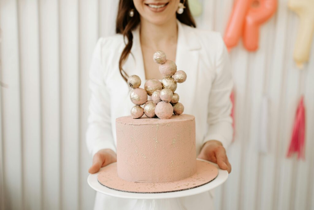 Pink cake with gold decorative balls held by woman in white blazer.